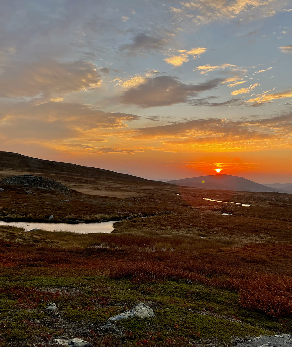 A gently sloping mountainside with a stream and the midnight sun in the background, standing over a hill.