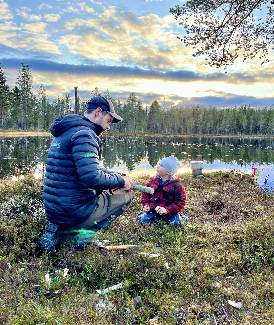 Raphael and Joël sitting on the ground by a lake while Raphael saws through a piece of wood.