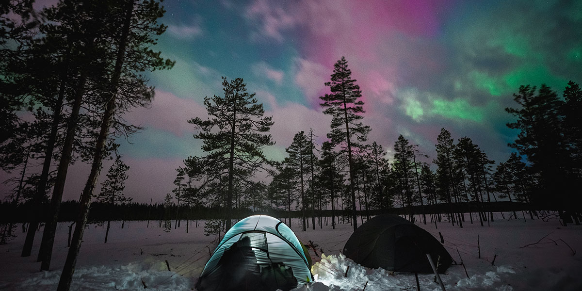 Two tents in the snow, under a lightly clouded sky illuminated by the northern lights.